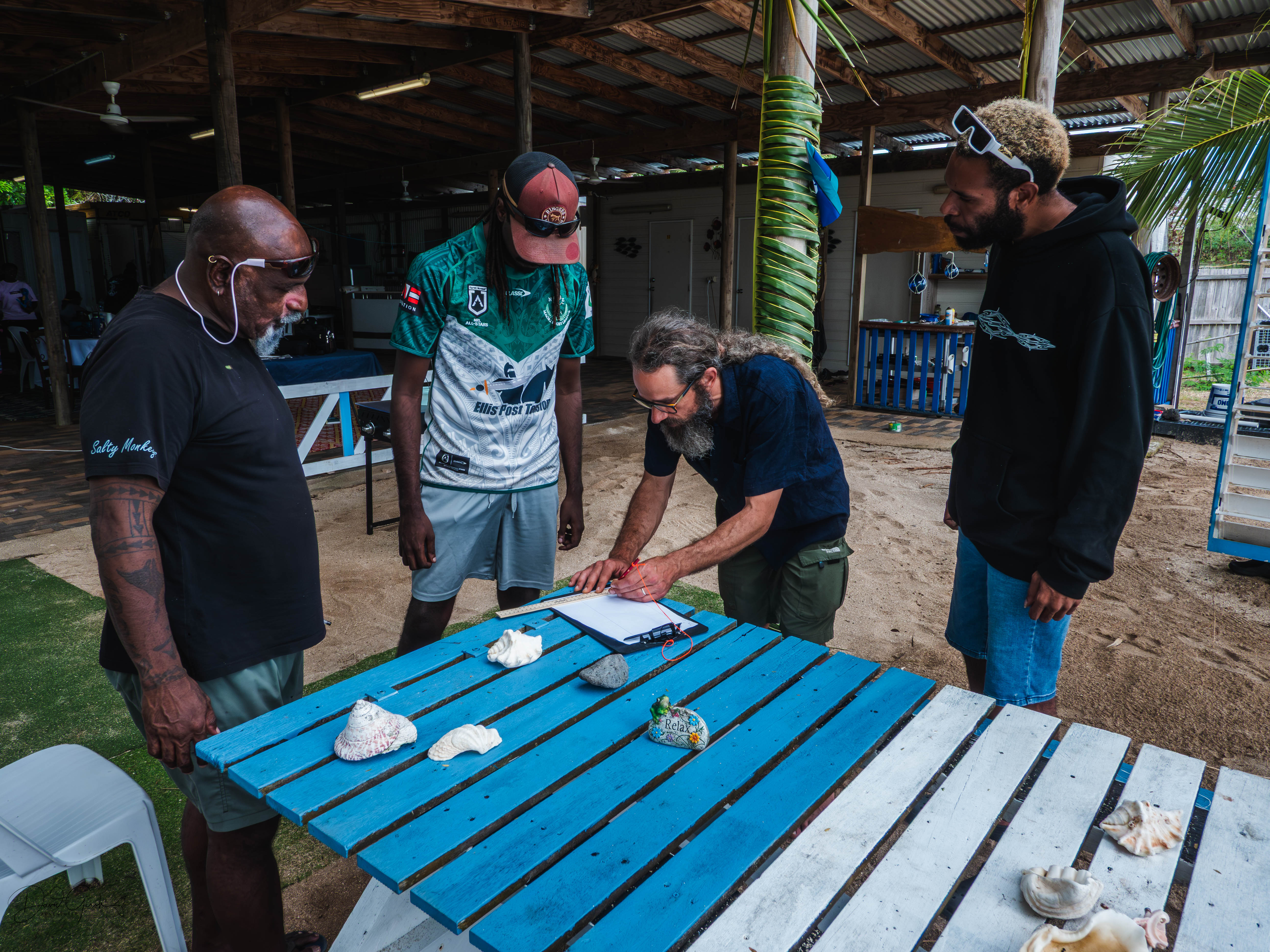 Men gathered around a table examining shells and stones on Mer Island